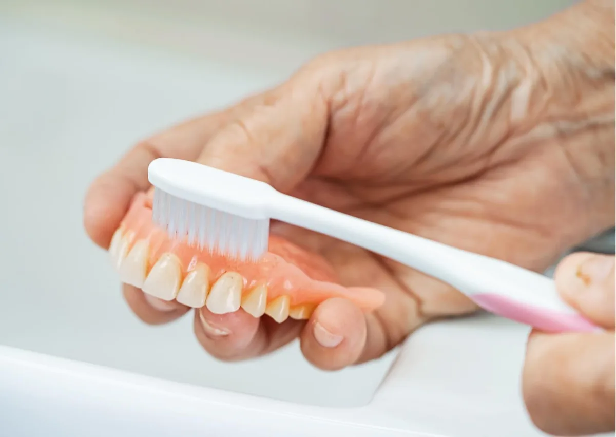 Toothbrush cleaning a removable denture as part of daily care when comparing dentures vs. implants.